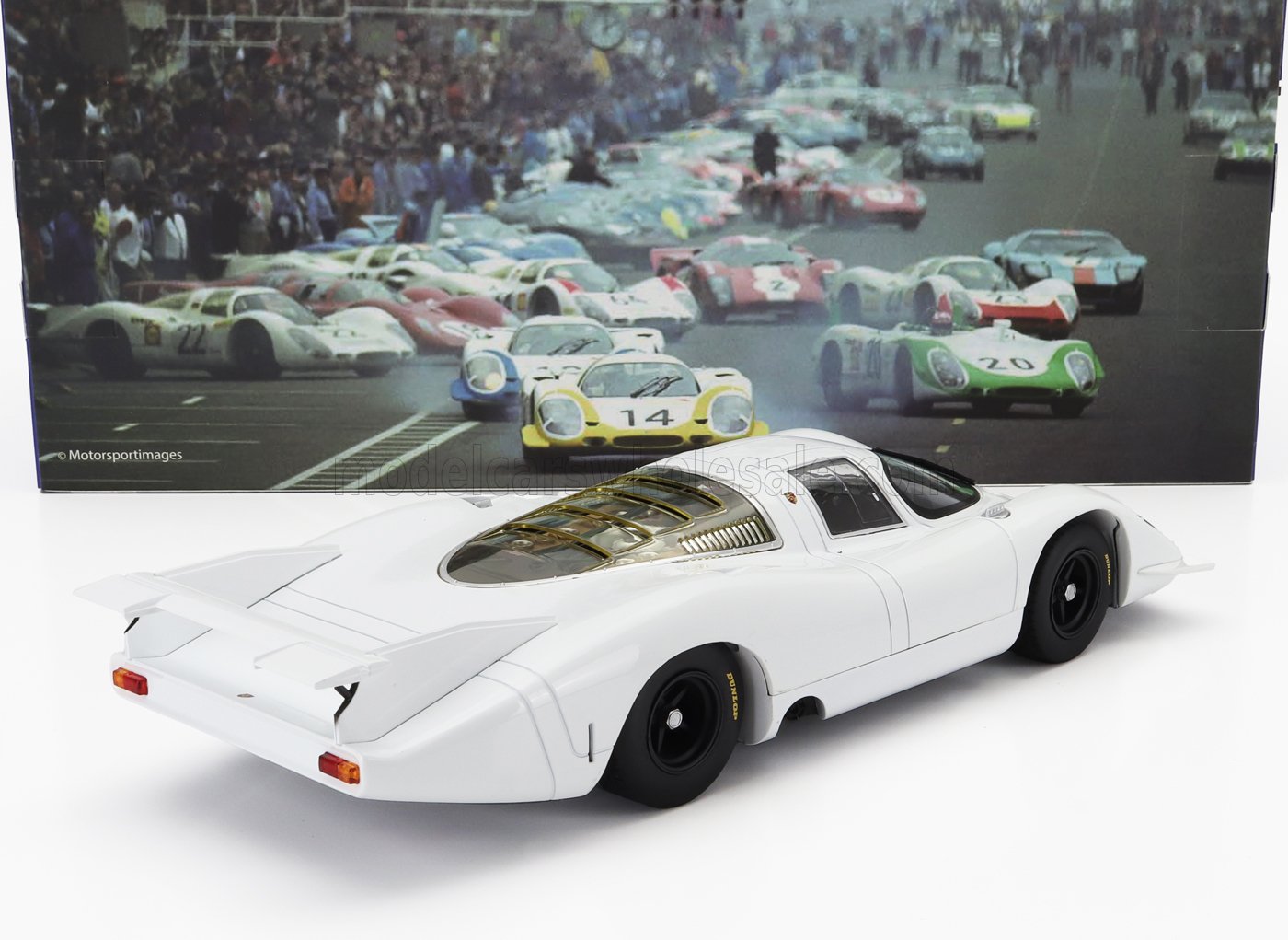 White vintage race car model with rear wing in front of classic racing poster, available on Vroomi
