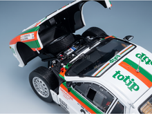 A detailed model of a white rally car with its hood open, featuring green and orange totip sponsor decals and a spare tire mounted in the open hood, photographed on a light gray studio background.