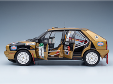 A scale model of a Lancia Delta Esso rally car of Tabaton-Tedeschini shown in side profile with its doors open. The car features a metallic gold and black livery with white wheels and various racing decals, including Esso and Mobil 1.