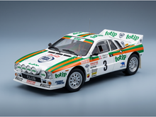 A scale model of a white Lancia rally car with green and orange stripes, featuring various sponsor logos including Totip, Pirelli, and Shell, set against a clean gray studio background.