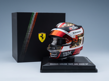 A miniature Charles Leclerc 2025 Abu Dhabi GP Ferrari helmet display on a black base, positioned in front of its black presentation box featuring the Ferrari logo.