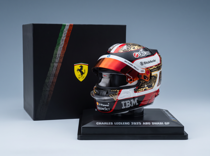 A miniature Charles Leclerc 2025 Abu Dhabi GP Ferrari helmet display on a black base, positioned in front of its black presentation box featuring the Ferrari logo.