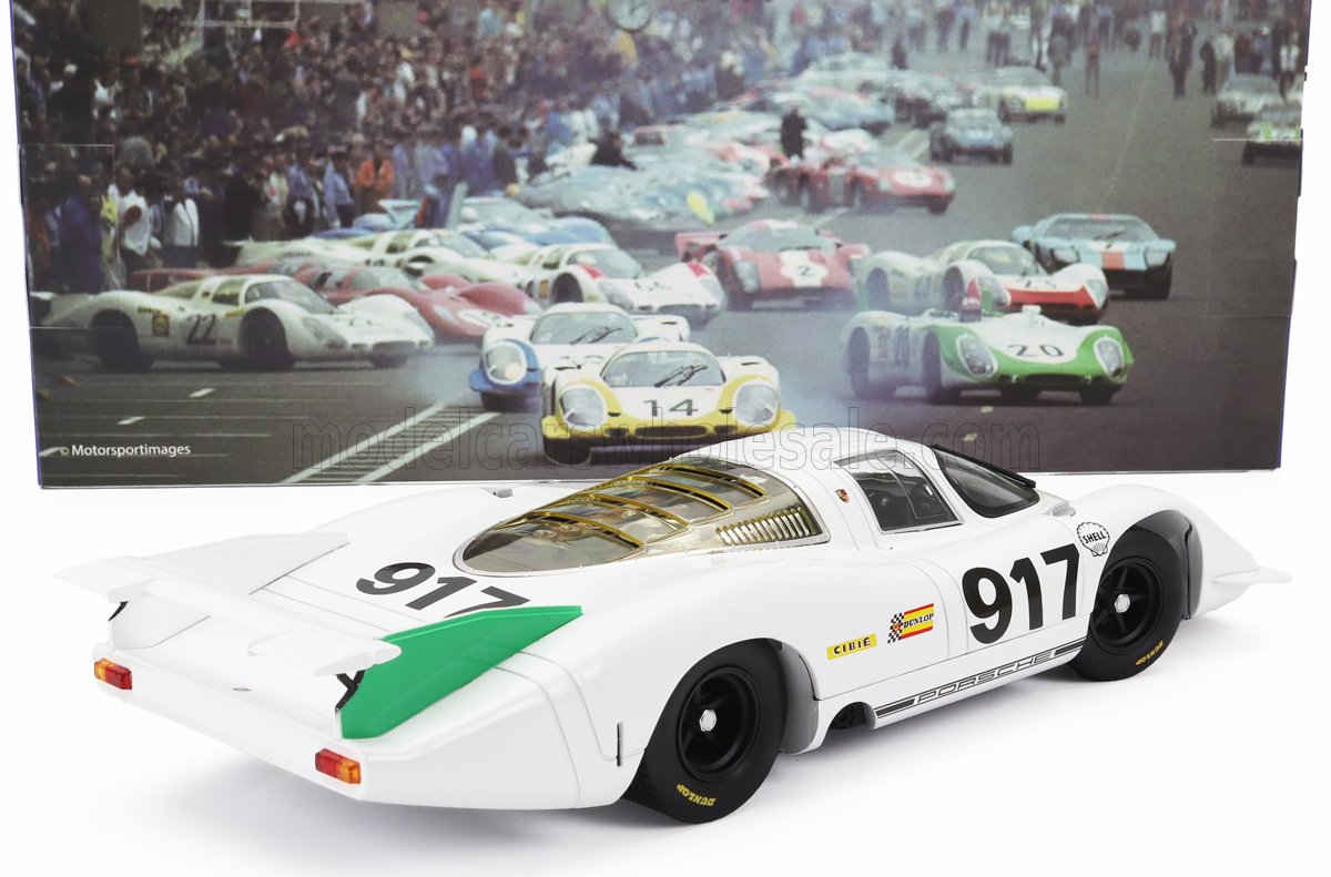 Modelo de coche de carreras Porsche 917 blanco con detalles en verde, expuesto delante de una foto clásica de las carreras de Le Mans, ahora a la venta en Vroomi.