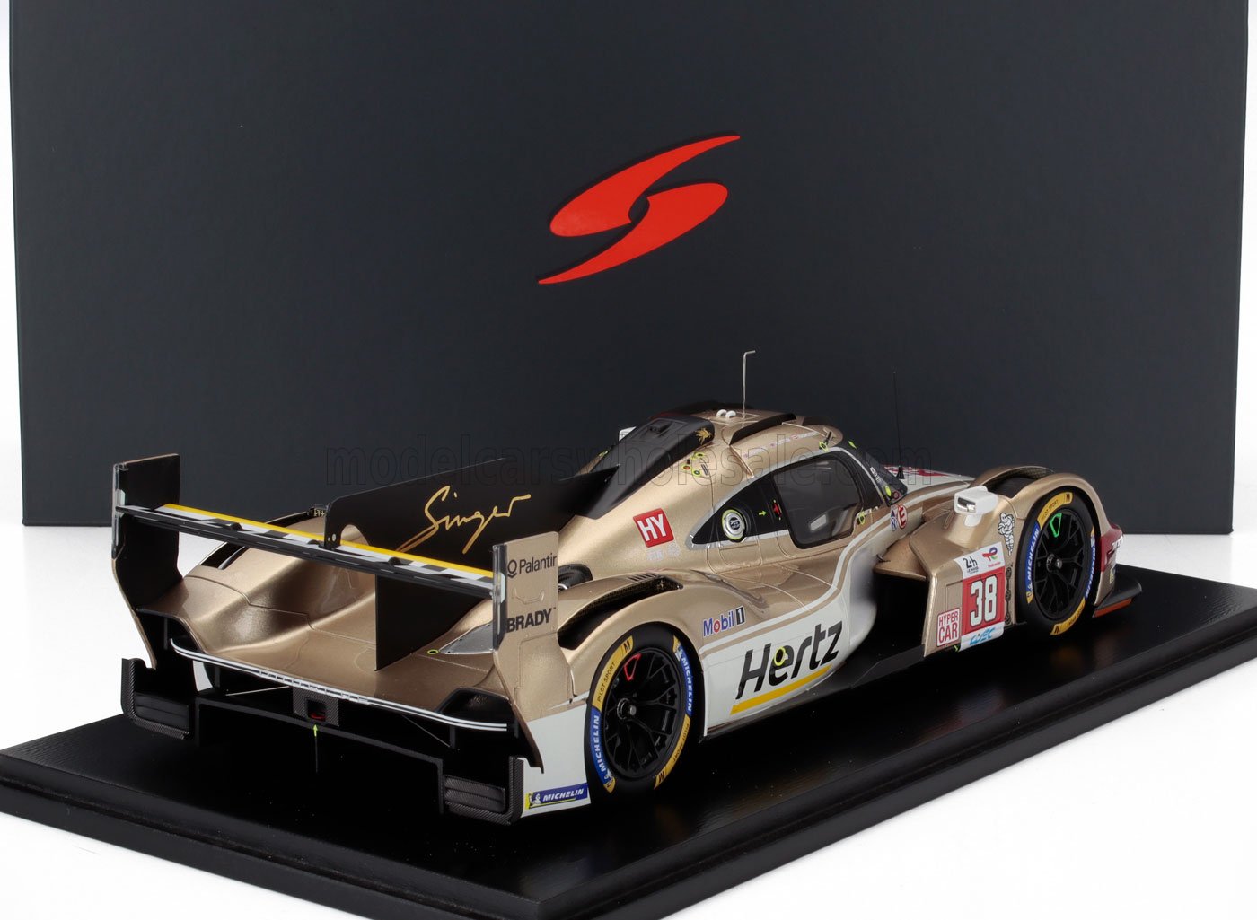 Coche de carreras Porsche 963 dorado y blanco con la marca Hertz en una vitrina negra, disponible en Vroomi.