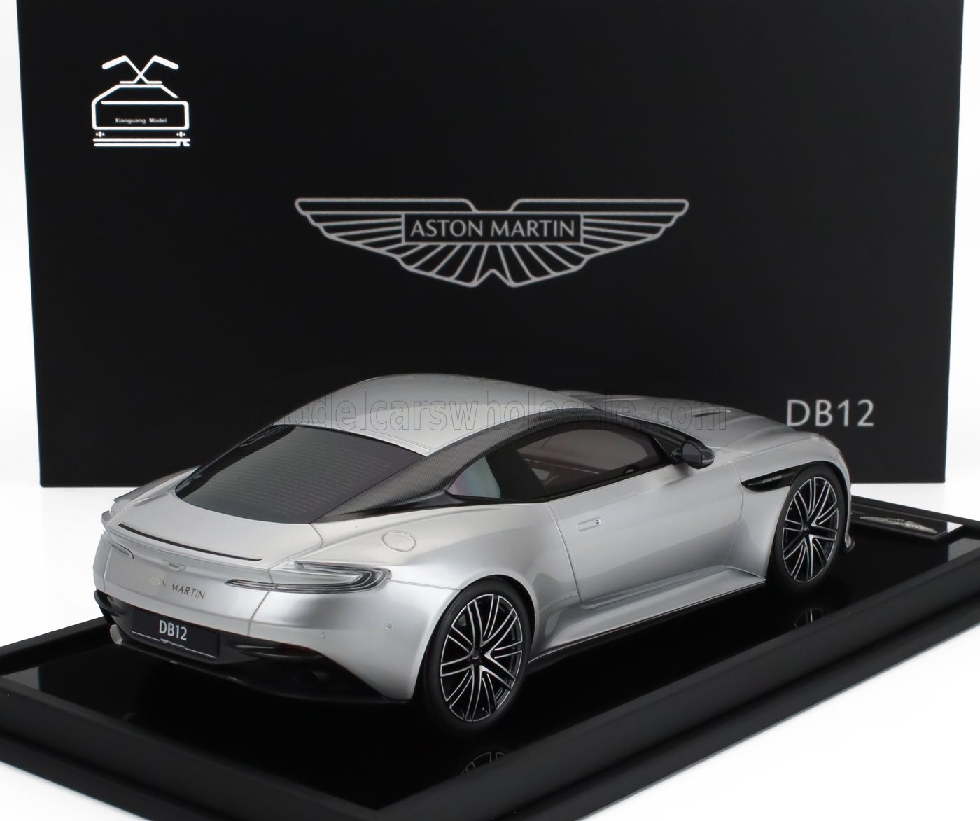 Modelo de coche Aston Martin DB12 plateado expuesto en un soporte negro con fondo de marca, disponible a través de Vroomi.