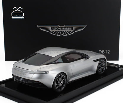 Modelo de coche Aston Martin DB12 plateado expuesto en un soporte negro con fondo de marca, disponible a través de Vroomi.