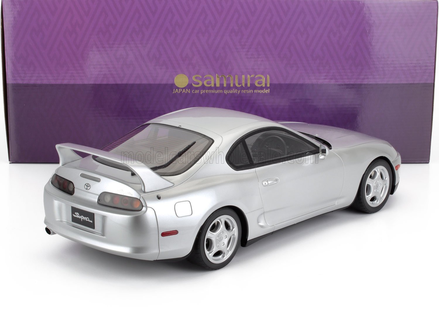 Modelo de coche Toyota Supra plateado con alerón trasero expuesto delante de una caja Samurai morada, también disponible en Vroomi.