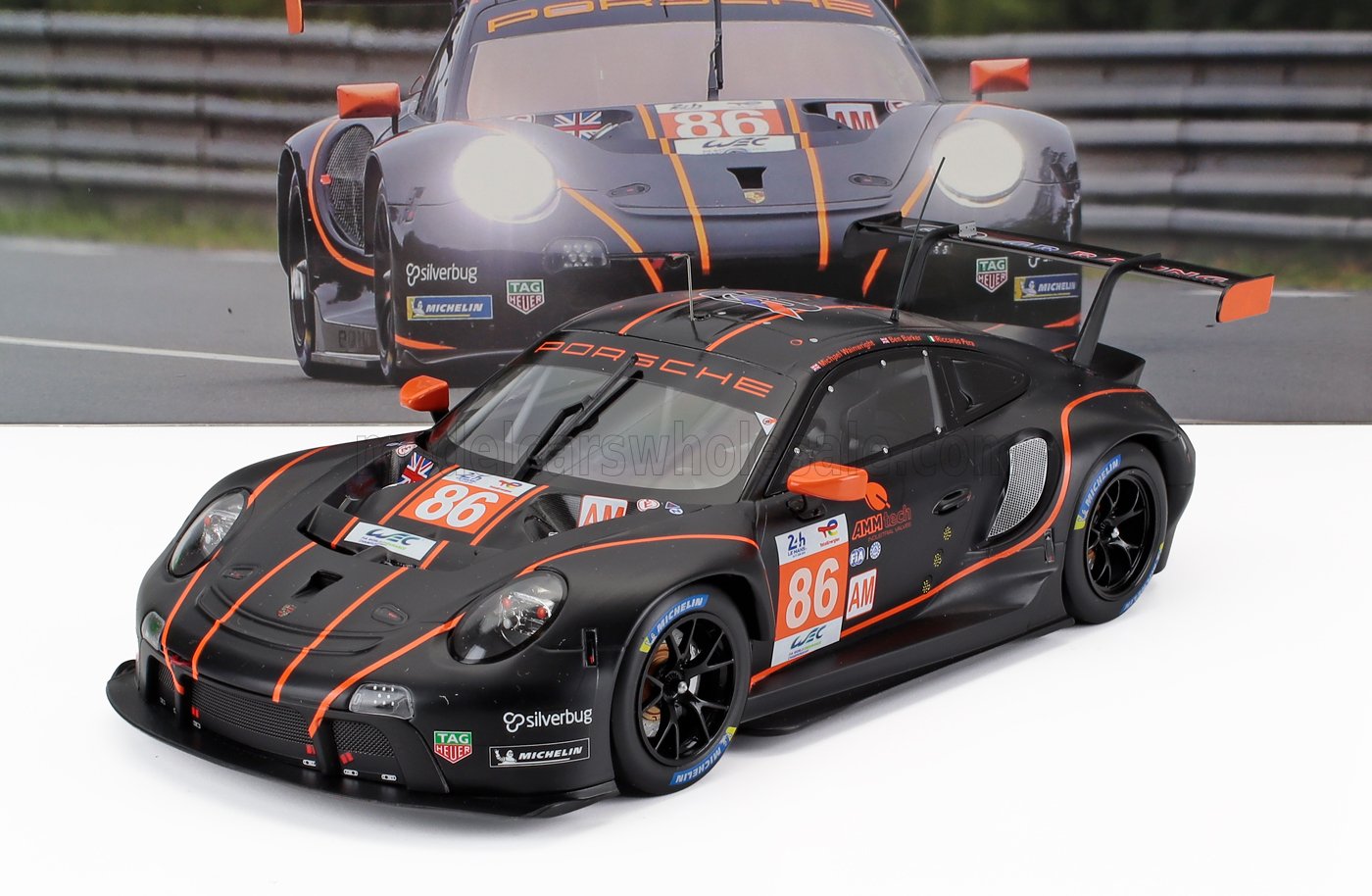 Matte black Porsche racing car model with orange stripes, number 86, and sponsor logos, showcased with race backdrop, now on Vroomi