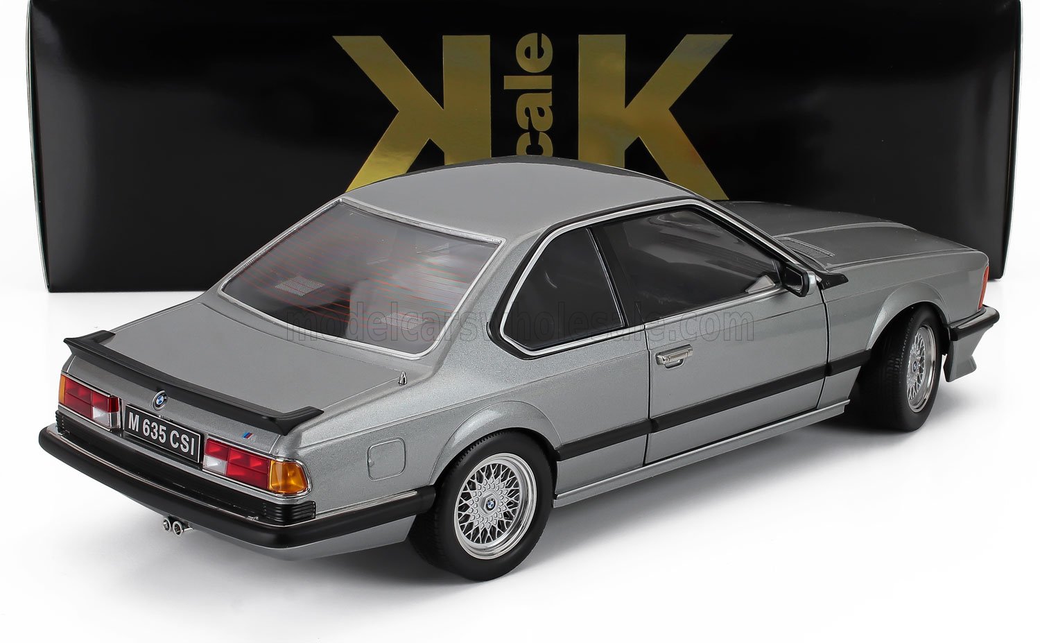 Modelo de coche BMW M635CSi plateado, vista trasera lateral con alerón negro y caja dorada a escala KK, disponible a través de Vroomi.