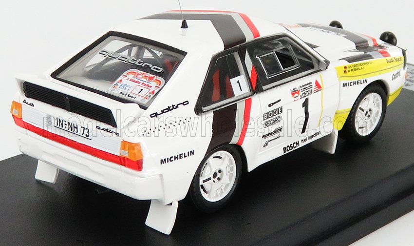 Modèle réduit de voiture de rallye Audi Quattro blanche avec décalcomanies de course et sponsors sur socle noir, disponible sur Vroomi.