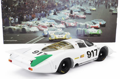 Modèle réduit de voiture de course Porsche 917 blanche avec des accents verts, exposé devant une photo classique des courses du Mans, désormais disponible sur Vroomi.