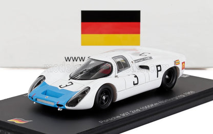 Modèle réduit Porsche 907 blanc et bleu sur socle d'exposition avec drapeau allemand en arrière-plan, proposé sur Vroomi.