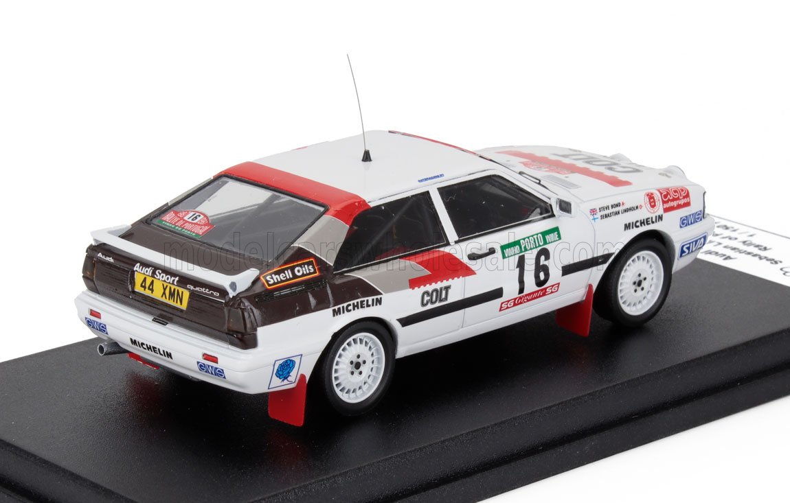 Modèle détaillé d'une voiture de rallye Audi Sport Quattro blanche avec des accents rouges et noirs, numéro 16, présenté sur un socle noir, proposé sur Vroomi.