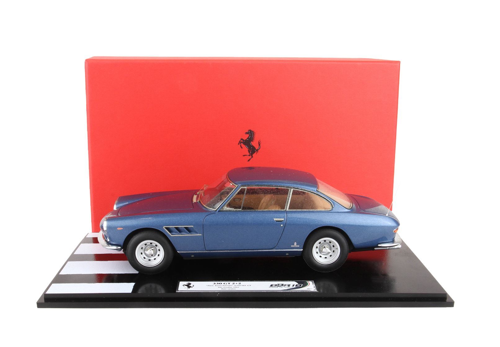 Blue Ferrari 330 GT 2+2 model car on display stand with red Ferrari backdrop, available on Vroomi