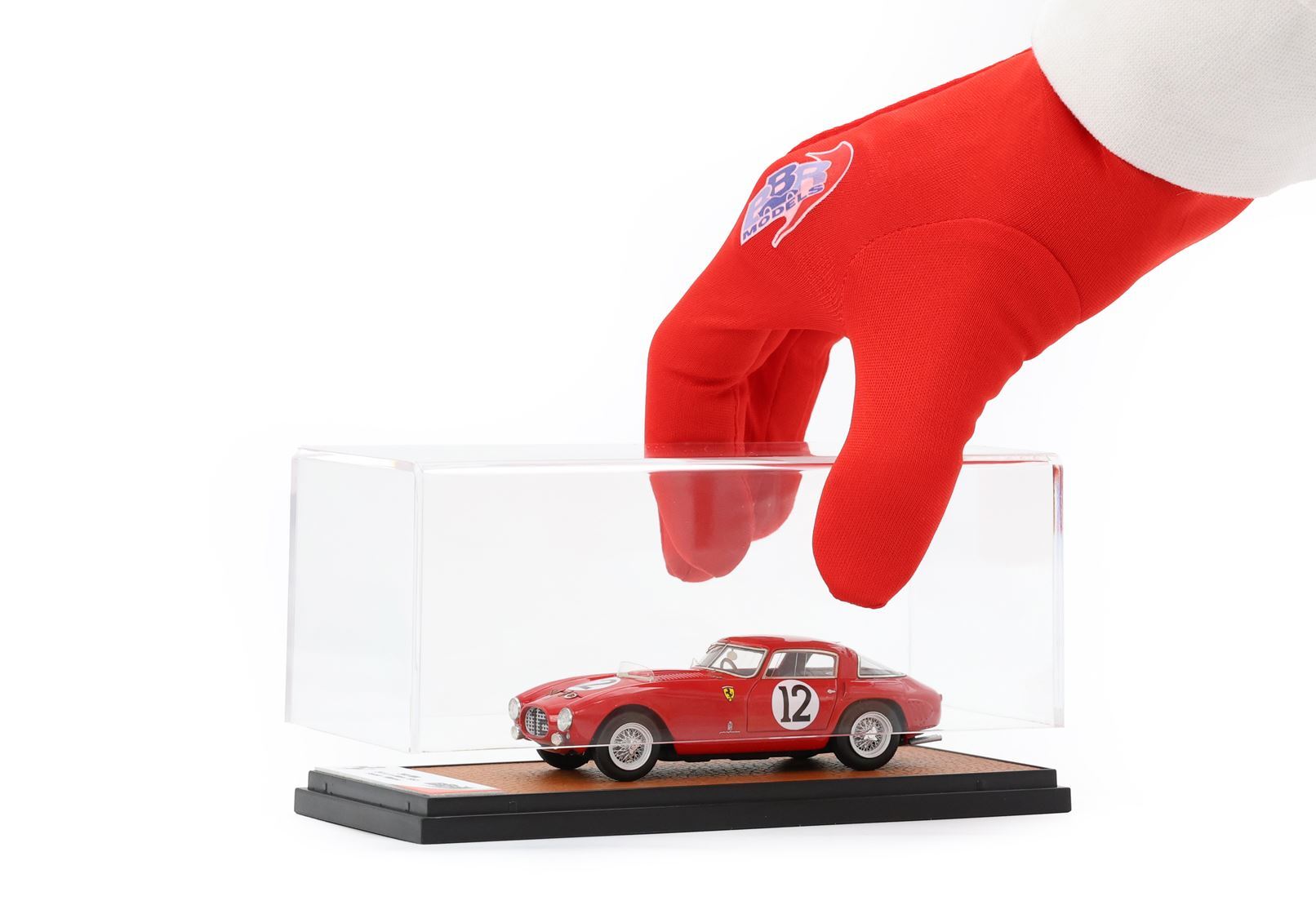 Main portant un gant rouge soulevant un étui transparent contenant une maquette rouge de la Ferrari numéro 12 sur un socle d'exposition, disponible via Vroomi.