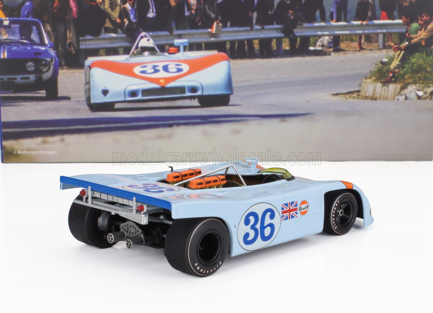 Modellino di auto da corsa blu con il numero 36, il logo Gulf e la bandiera britannica, mostrato con una foto vintage di una gara, disponibile su Vroomi.