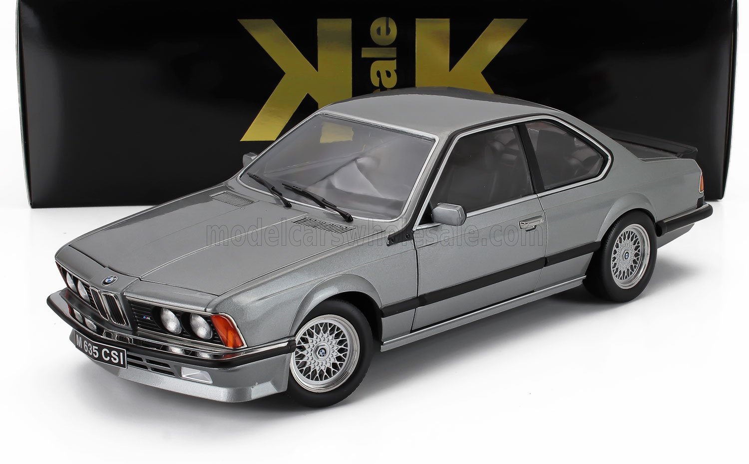 Modellino di BMW 635 CSi grigio con cerchi a razze, esposto davanti a una confezione nera e dorata, disponibile su Vroomi.