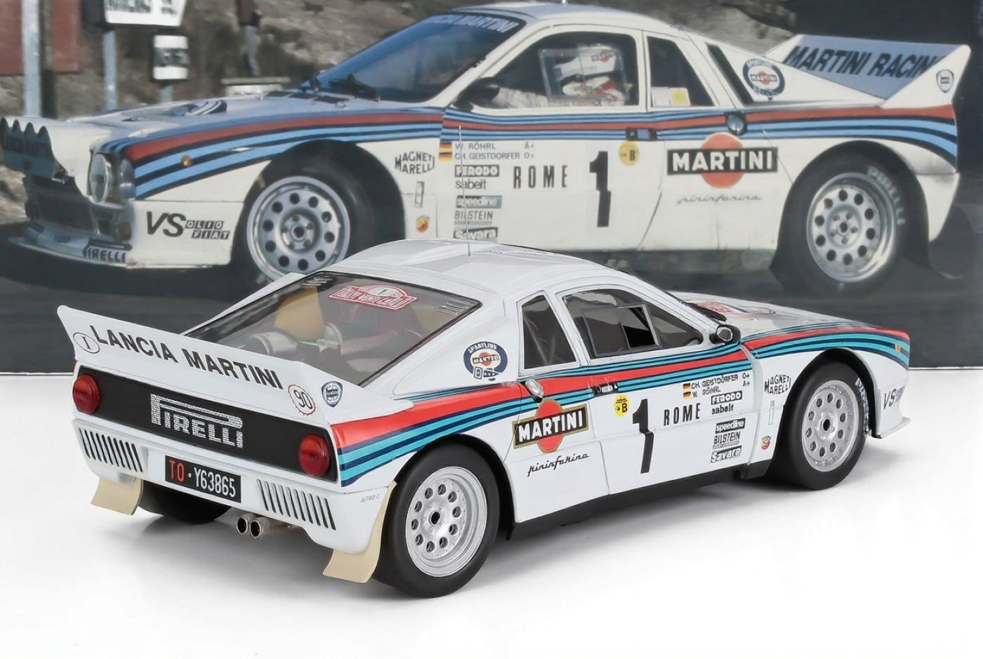 Modellino di auto da rally Lancia Martini bianca con adesivi degli sponsor, pneumatici Pirelli, mostrato su uno sfondo con foto di gara, presentato su Vroomi.